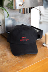 Schwarze Wien Streetwear Dad Cap mit roter Cafe Bauchstich Stickerei vorne, auf Holztisch vor Espressomaschine