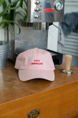 Rosa Dad Cap mit roter Cafe Bauchstich Stickerei, Wien Streetwear, auf Holztisch vor Siebträgermaschine